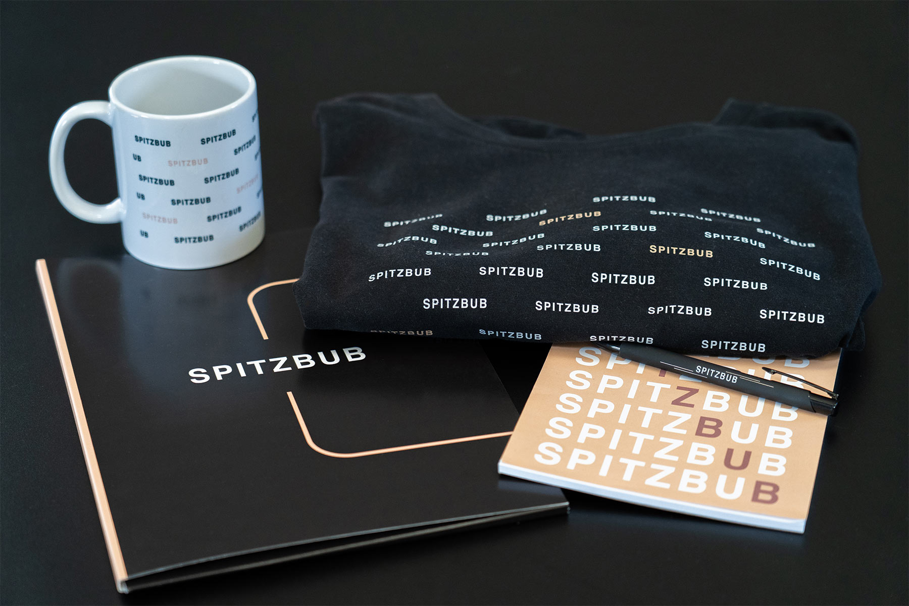 Eine Auswahl an schwarzen und weißen Merchandise-Artikeln der Marke „SPITZBUB“, darunter eine Tasse, ein gefalteter Pullover, ein Notizbuch mit Kugelschreiber und eine Mappe, die auf einer dunklen Oberfläche arrangiert sind.