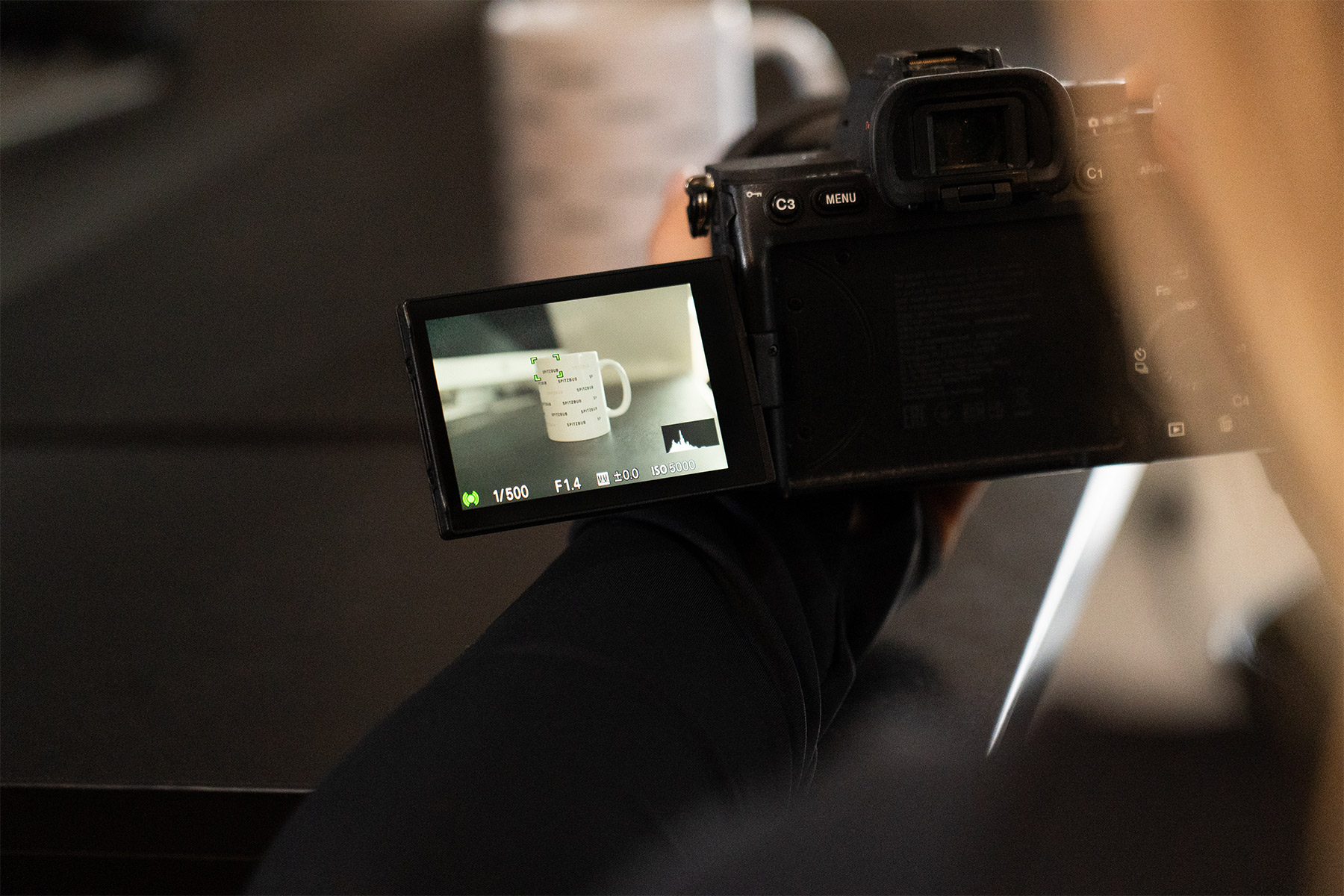 Eine Person fotografiert mit einer professionellen Kamera eine weiße Tasse, deren Motiv auf dem ausgeklappten Kameradisplay deutlich zu sehen ist.